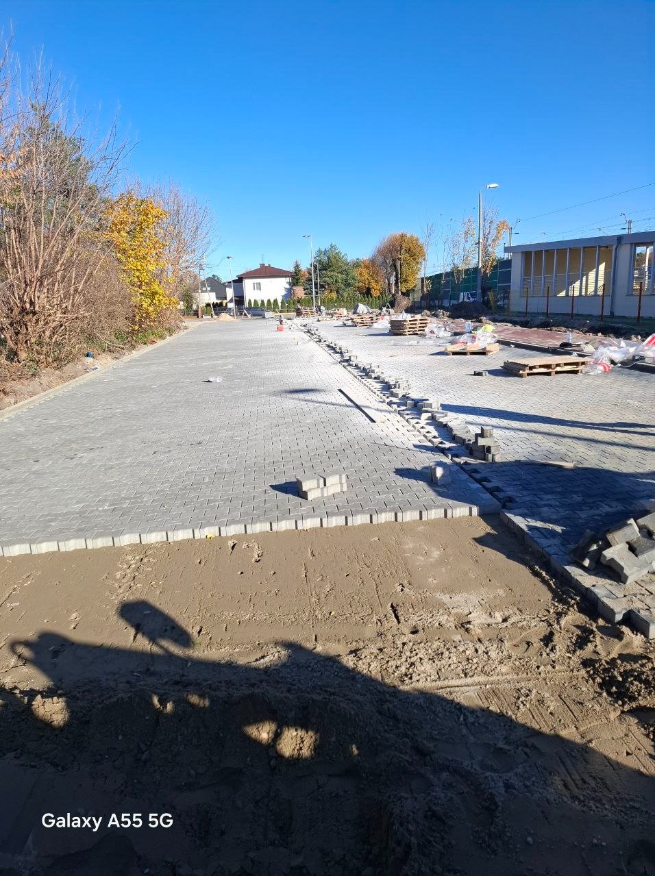 Obszar z częściowo ułożoną szarą kostką brukową, palety z kostką w tle, widoczne przygotowane podłoże z piasku, słoneczny dzień.