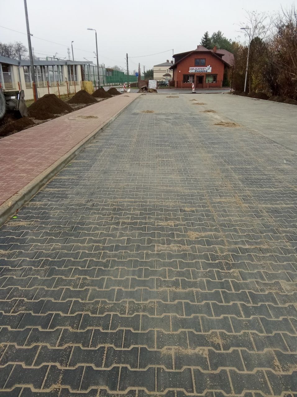 Nowa nawierzchnia z szarej kostki brukowej typu 'I', z widocznymi śladami piasku w spoinach, obok czerwonego chodnika i sterty ziemi, z budynkiem w tle.