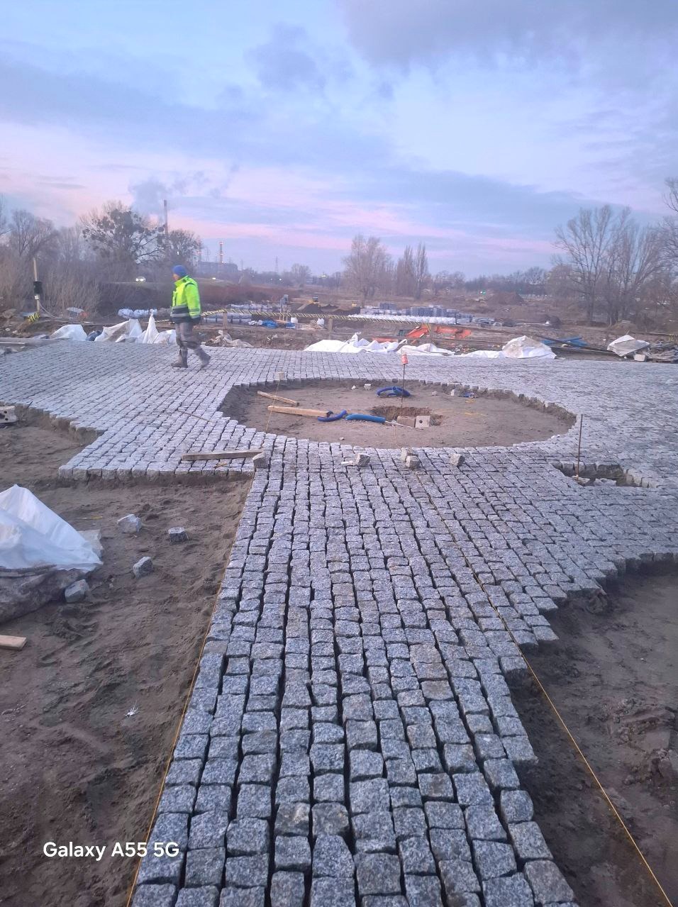 Obszar w trakcie wykańczania z szarej kostki granitowej, widoczny pracownik w odblaskowej kurtce, w tle elementy budowy i pochmurne niebo.