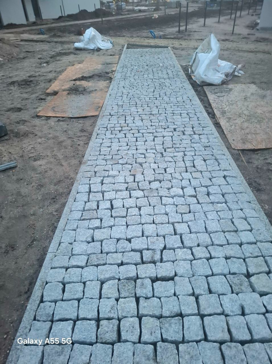 Świeżo ułożony chodnik z kostki granitowej o szarym odcieniu, z widocznymi śladami prac budowlanych i materiałami zabezpieczonymi folią w tle.