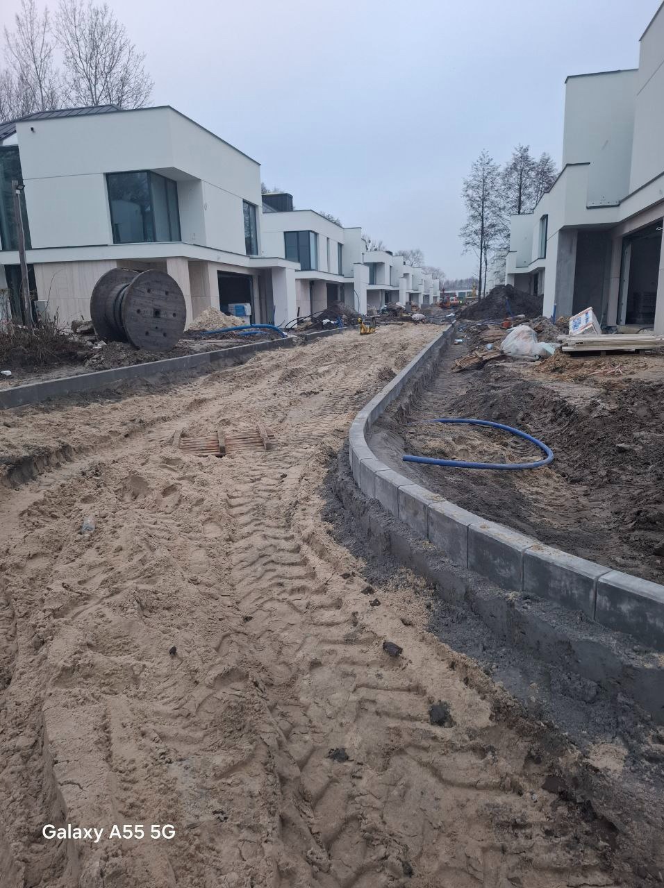 Ulica w trakcie budowy osiedla domów jednorodzinnych, widoczne krawężniki i piasek z wyraźnymi śladami opon maszyn budowlanych, w tle szpula z kablem.