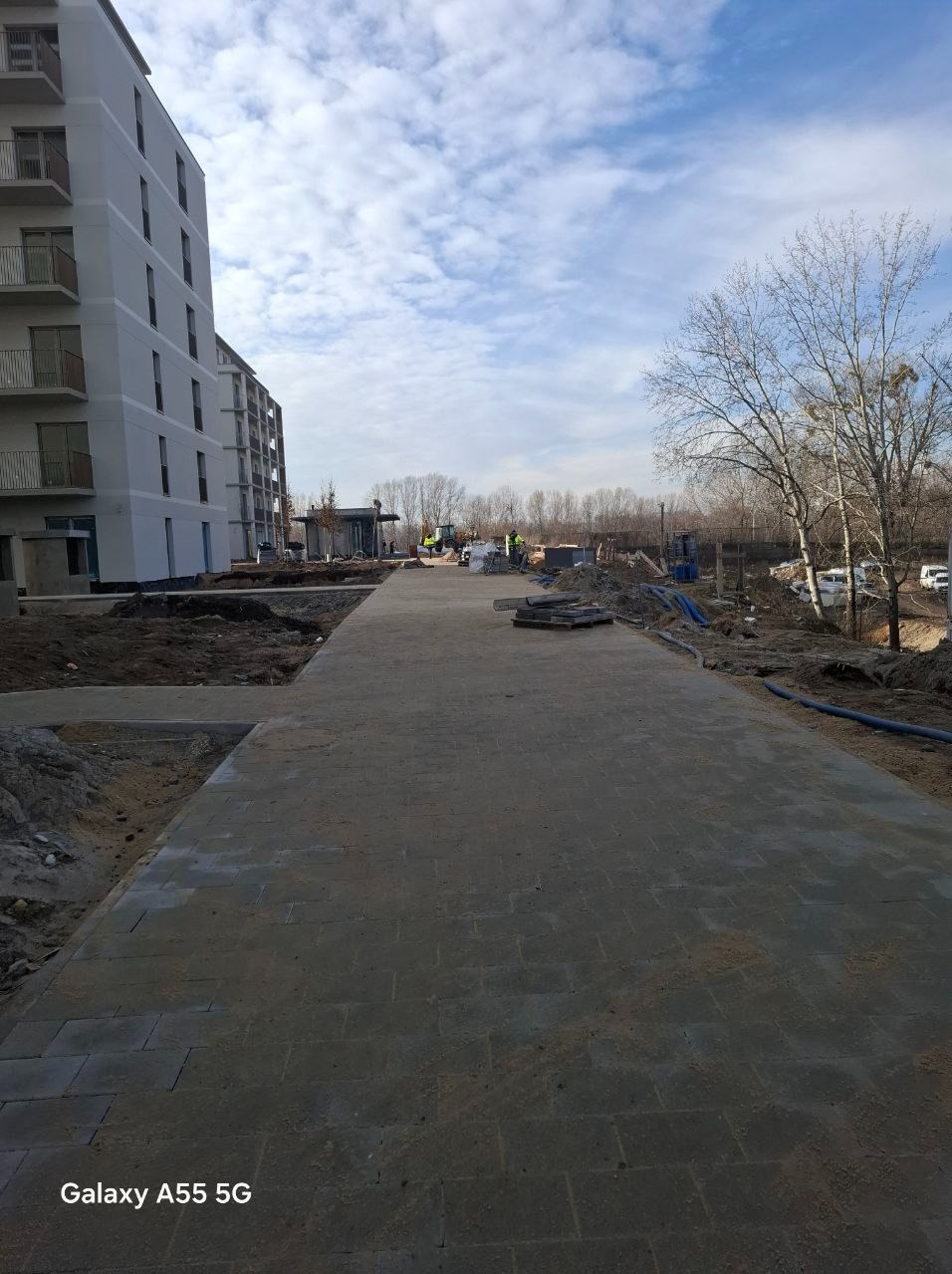 Szeroki chodnik z kostki brukowej prowadzący wzdłuż nowego budynku mieszkalnego, z widocznymi przygotowaniami do dalszych prac brukarskich i ziemnych w tle.