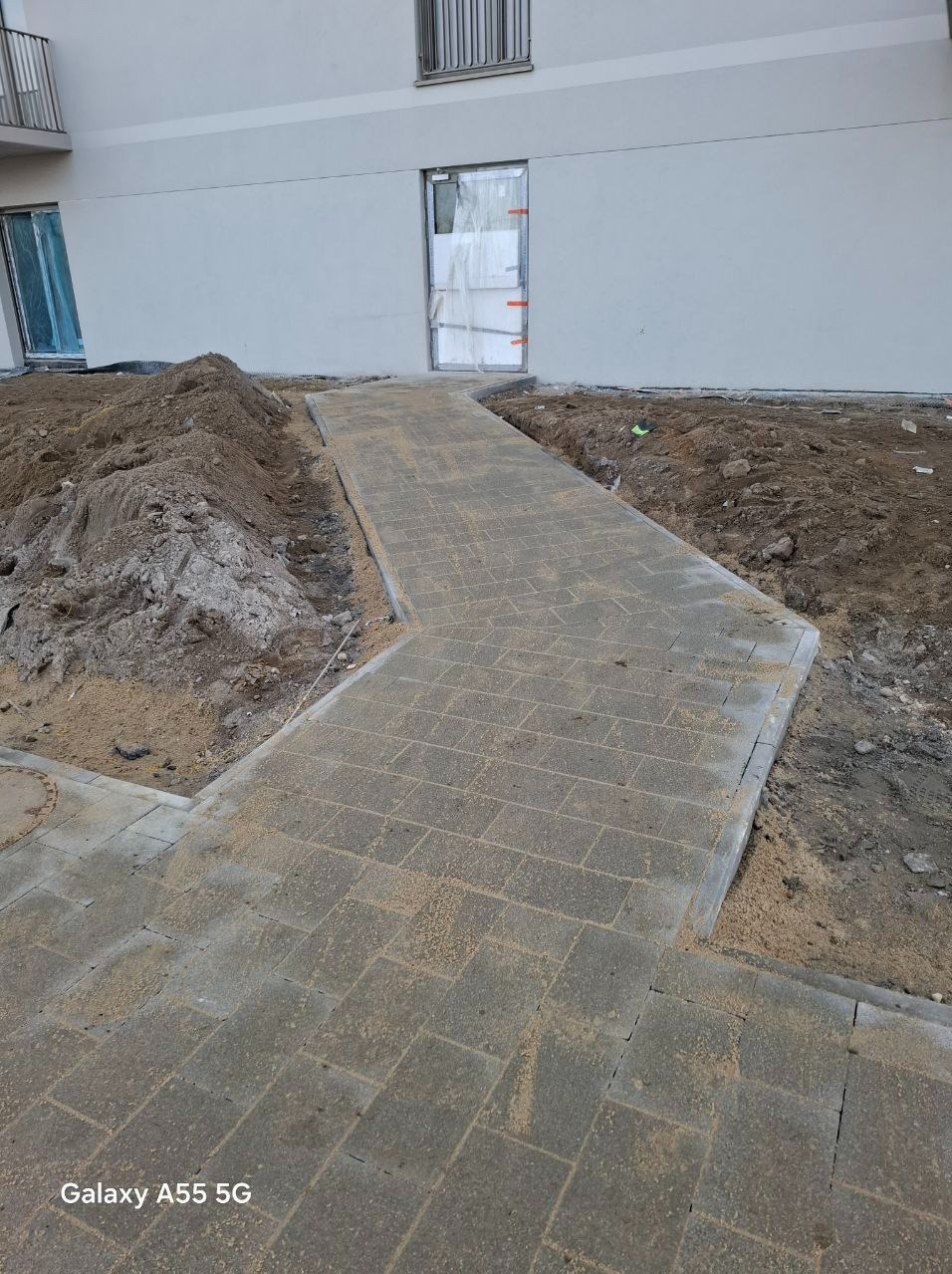Świeżo ułożona ścieżka z szarej kostki brukowej prowadząca do budynku, widoczny stos ziemi obok, piasek na kostce, częściowo ukończona praca.
