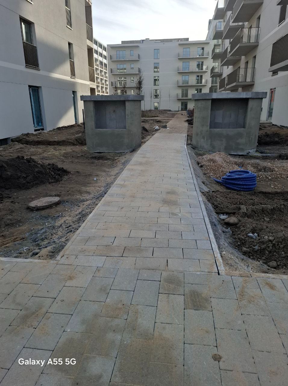 Ścieżka z jasnoszarej kostki brukowej prowadząca między dwoma betonowymi konstrukcjami w nowo powstającym osiedlu mieszkaniowym, obok widoczny zwój niebieskiego węża.