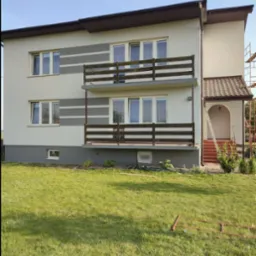 Dwupiętrowy dom z balkonami i świeżo odnowioną elewacją w kolorach beżu i szarości, widoczny z trawnika. Fragment rusztowania przy wejściu.