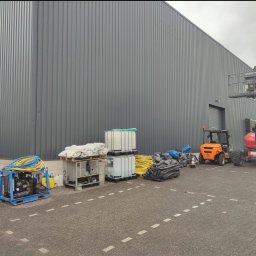 Festmal - Sprzęt do czyszczenia elewacji budynku przemysłowego: agregat ciśnieniowy, zbiorniki na wodę, worki z zabezpieczeniami i podnośniki koszowe.