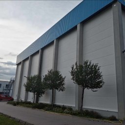 Festmal - Duży, biały magazyn z niebieskim dachem blaszany, otoczony trzema małymi drzewkami i chodnikiem z żółtym kablem na obrzeżach.