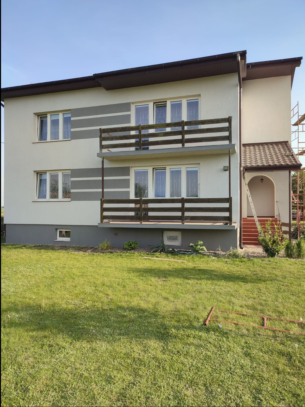 Dwupiętrowy dom z balkonami i świeżo odnowioną elewacją w kolorach beżu i szarości, widoczny z trawnika. Fragment rusztowania przy wejściu.