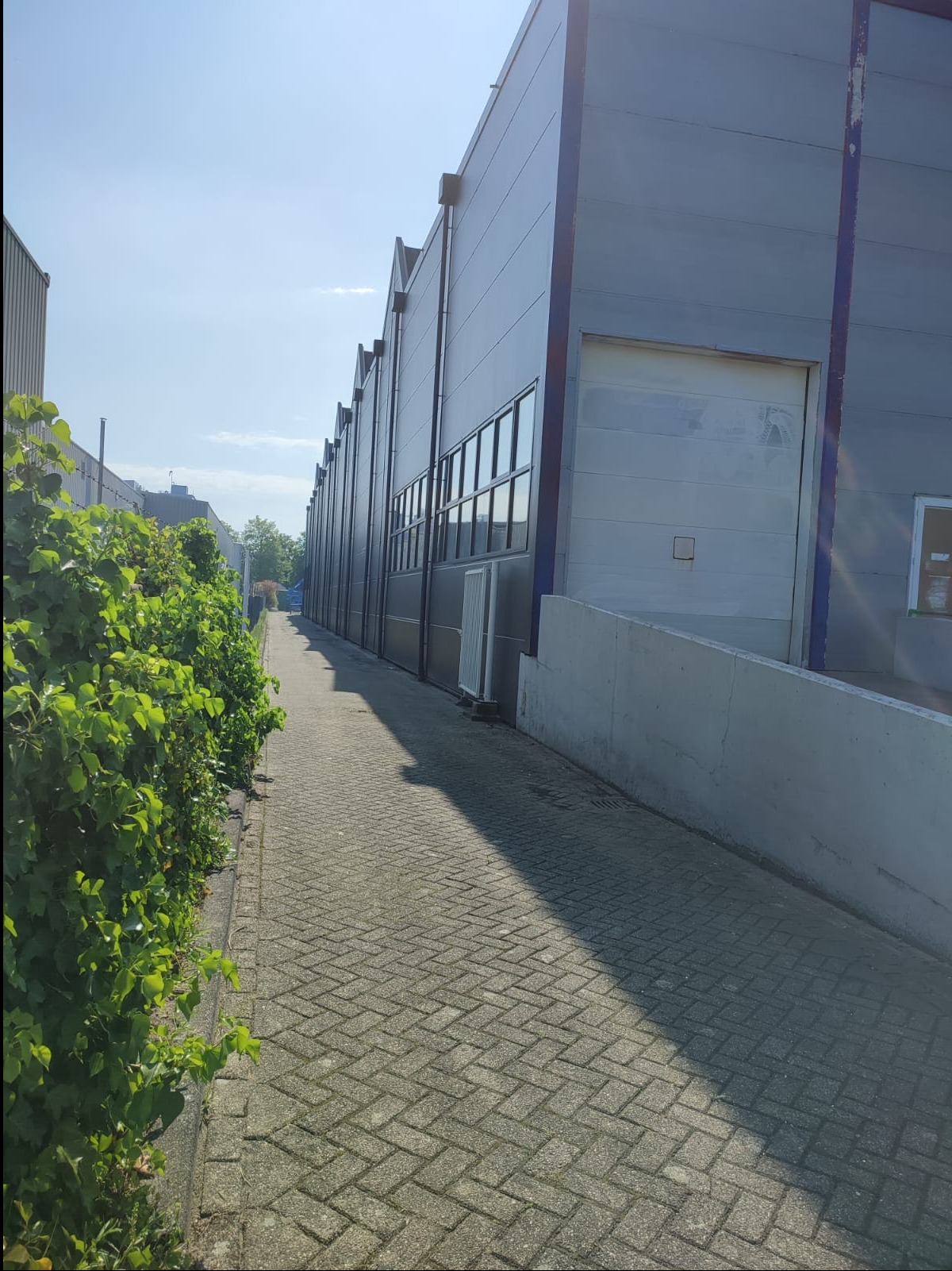 Długa, szara elewacja budynku przemysłowego z rzędem okien i bramą wjazdową, widoczna z perspektywy chodnika obok zielonego żywopłotu w słoneczny dzień.