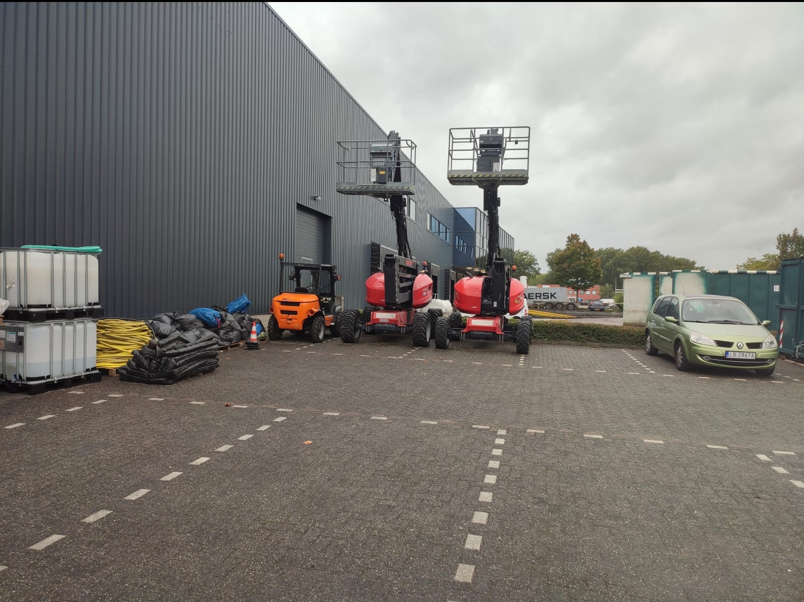 Dwie podnośniki koszowe na parkingu przed budynkiem o szarej elewacji, obok wózek widłowy i zaplecze budowlane