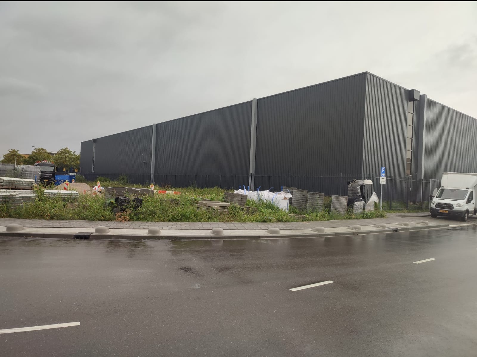 Długi, ciemnoszary budynek przemysłowy z blachy falistej, widoczny z mokrej ulicy; w tle zaparkowane pojazdy i składowane materiały budowlane.