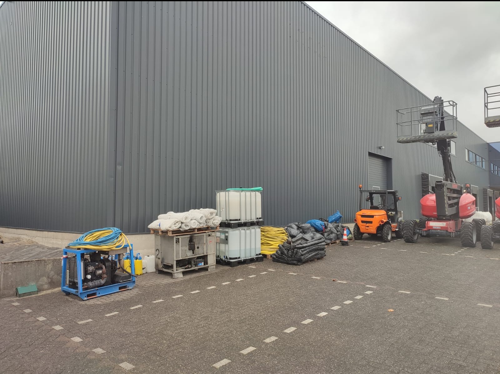 Sprzęt do czyszczenia elewacji budynku przemysłowego: agregat ciśnieniowy, zbiorniki na wodę, worki z zabezpieczeniami i podnośniki koszowe.