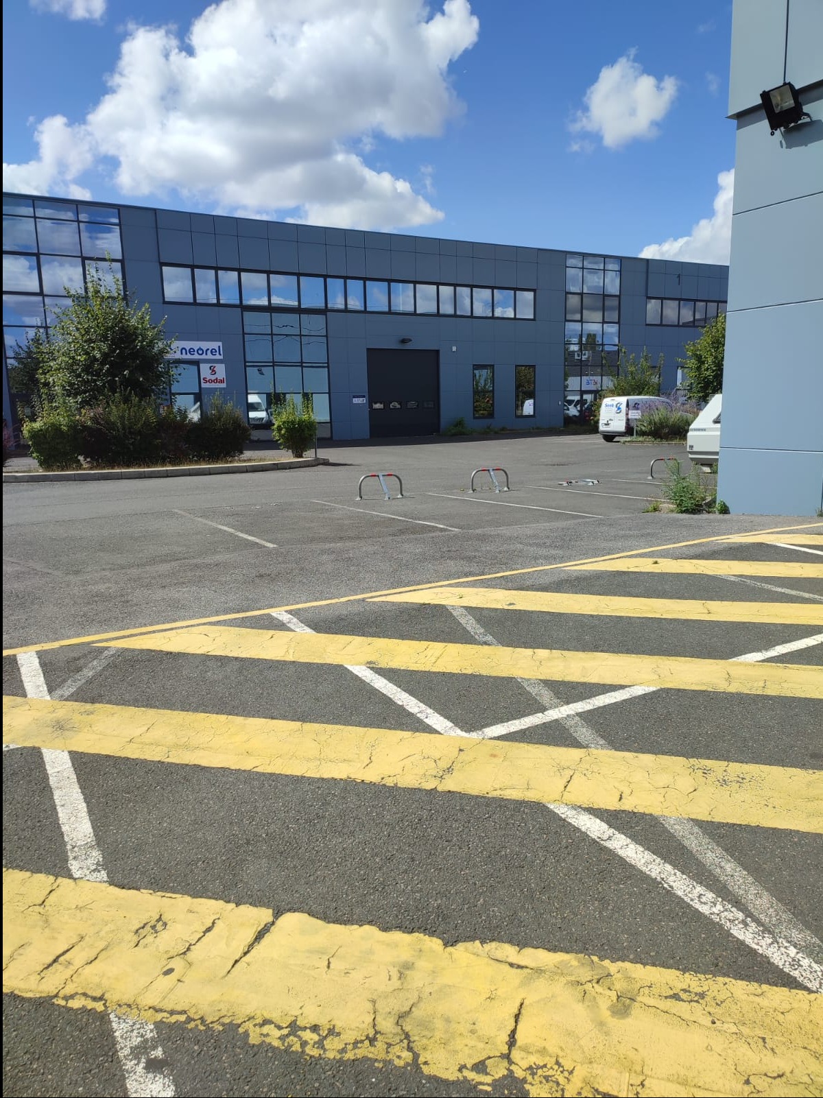 Szary budynek biurowy z rzędem okien, widoczny z parkingu z żółtymi pasami, w tle błękitne niebo z chmurami.