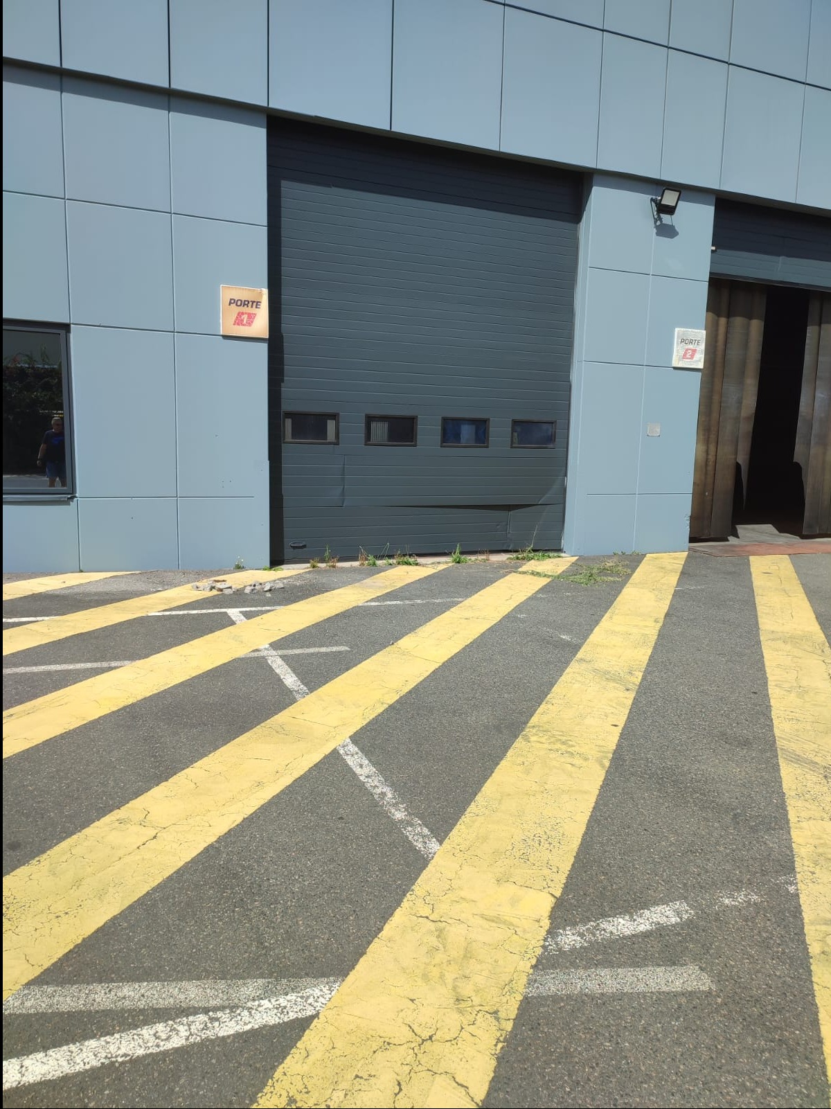 Szara fasada budynku przemysłowego z częściowo otwartą, segmentową bramą garażową z małymi oknami, żółte pasy na asfalcie przed budynkiem.