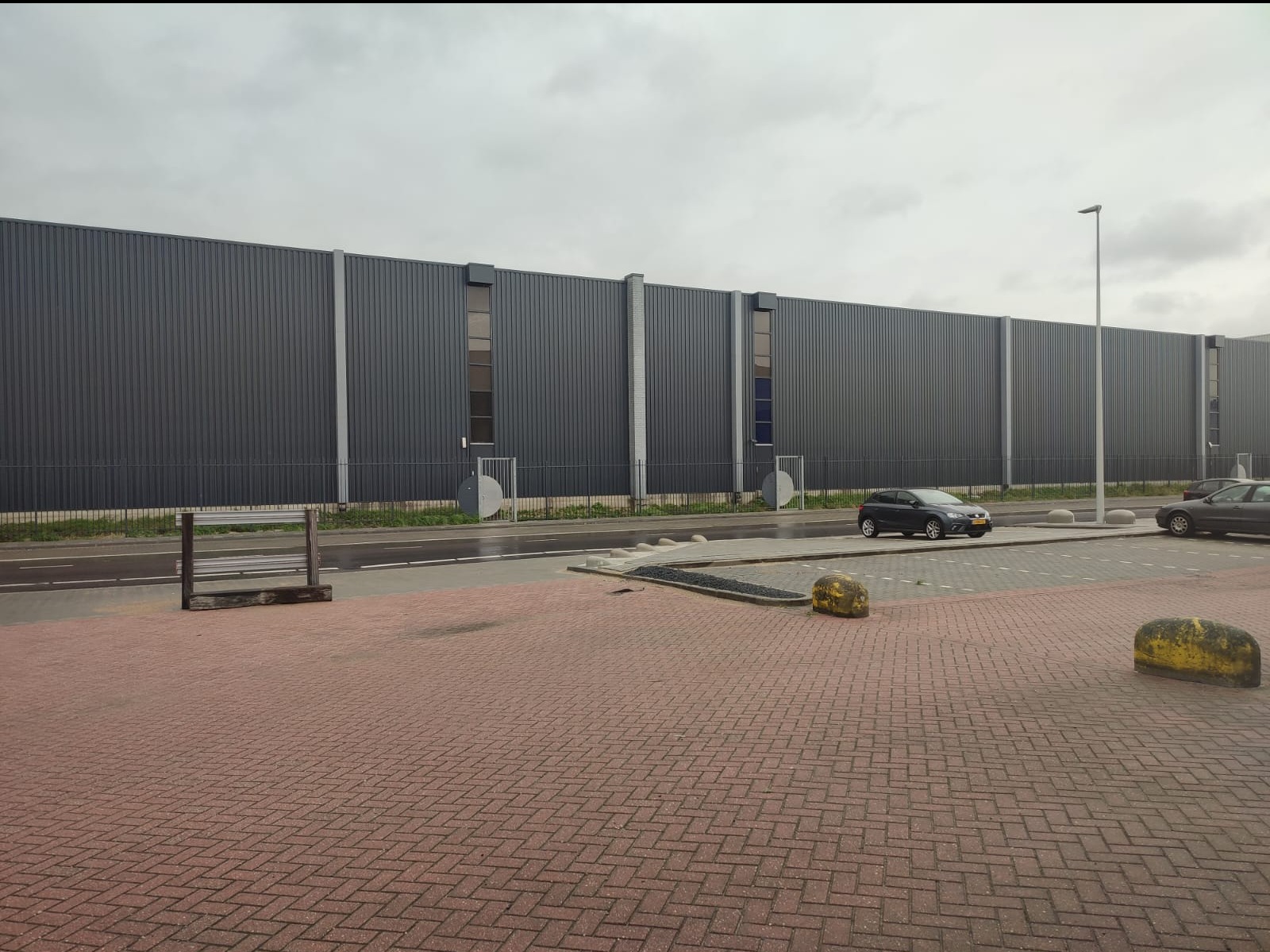 Długa, szara elewacja budynku przemysłowego z pionowymi panelami, widoczna z poziomu chodnika wyłożonego czerwoną kostką. Na pierwszym planie kamienne bariery i zaparkowane samochody.