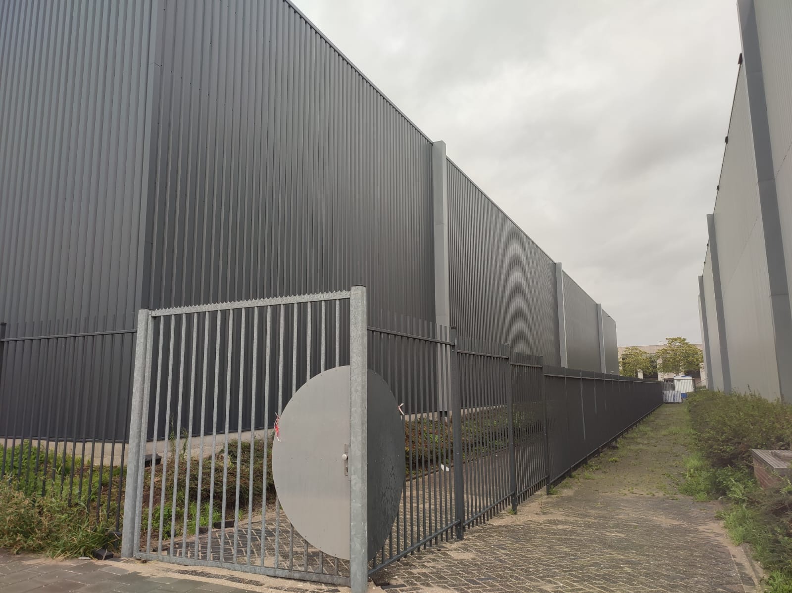 Szara, metalowa elewacja budynku przemysłowego z widoczną bramą wjazdową i ogrodzeniem z metalowych prętów.