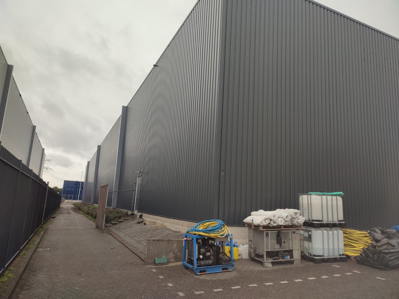 Szara, metalowa elewacja budynku przemysłowego z widocznym sprzętem do czyszczenia ciśnieniowego, w tym agregat z niebieską ramą i zwinięte węże, paleta z białymi workami i zbiorniki na wodę,...