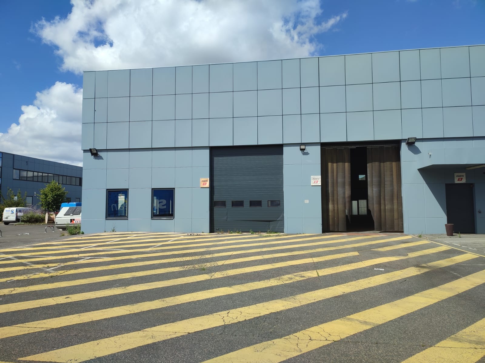 Szary budynek przemysłowy z panelową elewacją, otwartą bramą rolowaną i oknami, z żółtymi pasami na podłożu.