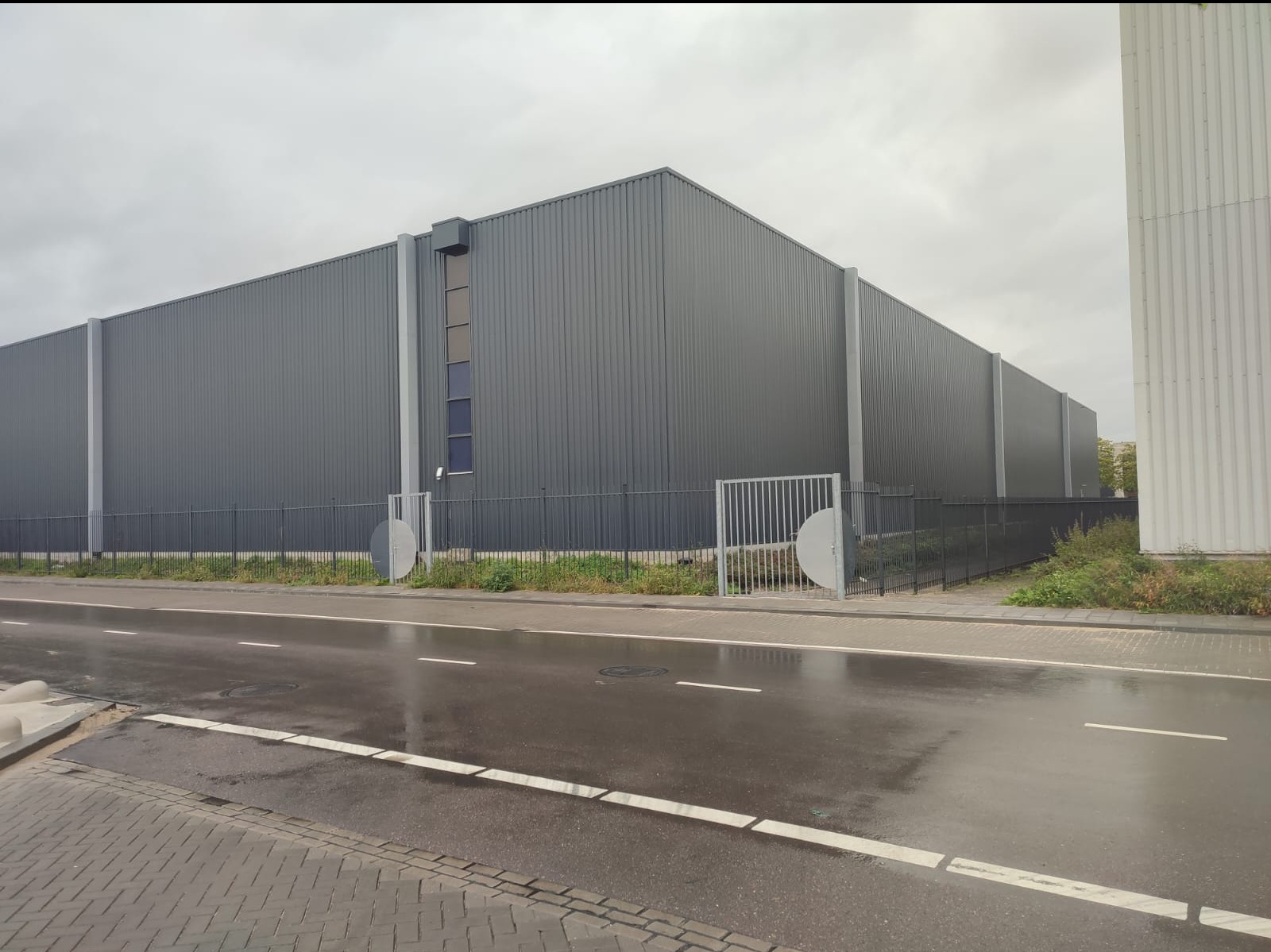 Duży, szary budynek przemysłowy z blachy trapezowej, widoczny z poziomu ulicy po deszczu, z metalowym ogrodzeniem i bramą w tle pochmurnego nieba.