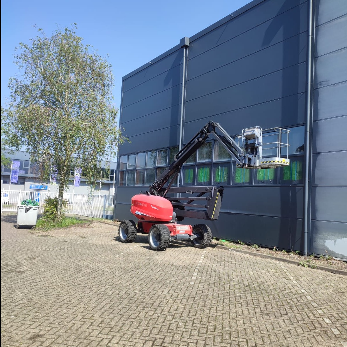 Czerwony podnośnik koszowy ustawiony przed budynkiem o ciemnoszarej elewacji, widoczne okna zabezpieczone folią ochronną, przygotowanie do prac na wysokościach.