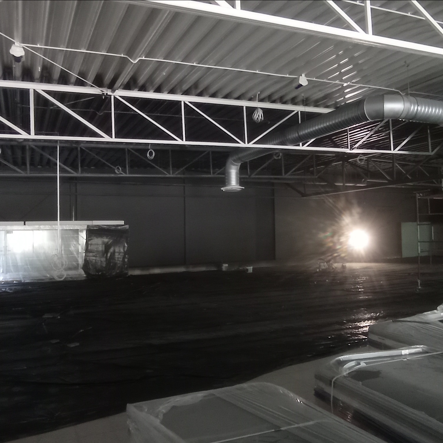 Przygotowanie hali produkcyjnej do malowania: podłoga zabezpieczona czarną folią, widoczne elementy konstrukcji dachu i wentylacji, pakunki materiałów budowlanych w folii czekające na użycie.