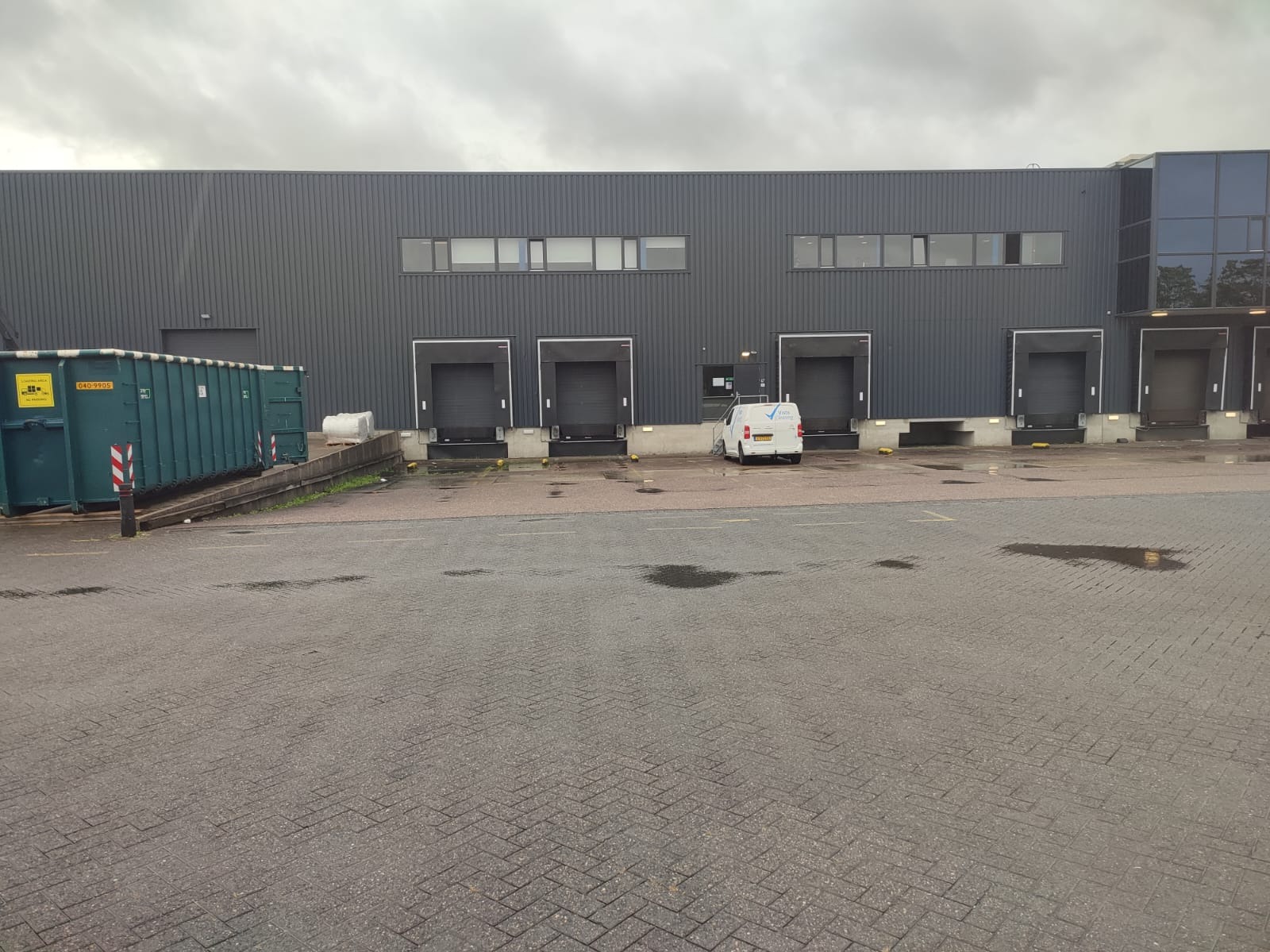 Szara hala magazynowa z czterema bramami załadunkowymi, kontenerem na śmieci po lewej i zaparkowanym białym vanem z logo firmy przed jedną z bram.