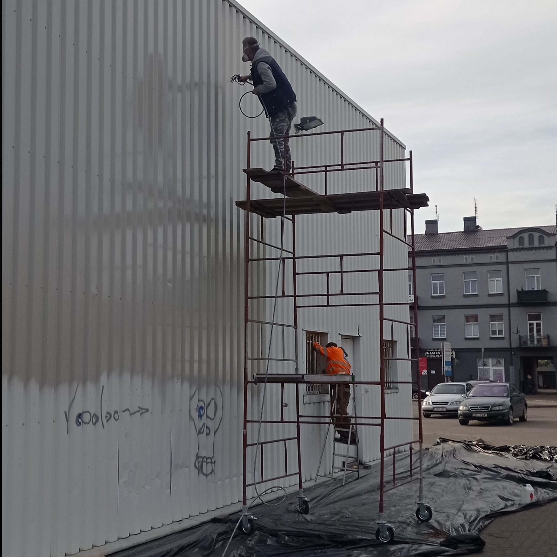 Pracownicy na rusztowaniu natryskowo pokrywają farbą blaszane ściany budynku, na których widoczne są graffiti.