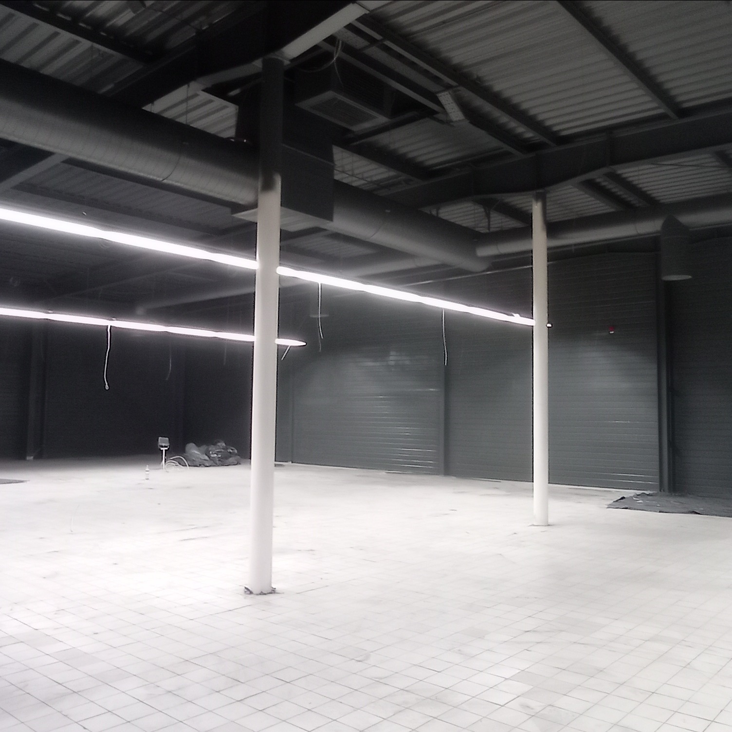 Wnętrze hali magazynowej z szarymi ścianami z blachy falistej, białymi kolumnami, szarą konstrukcją dachu i rurami wentylacyjnymi, oświetlone białymi lampami jarzeniowymi.