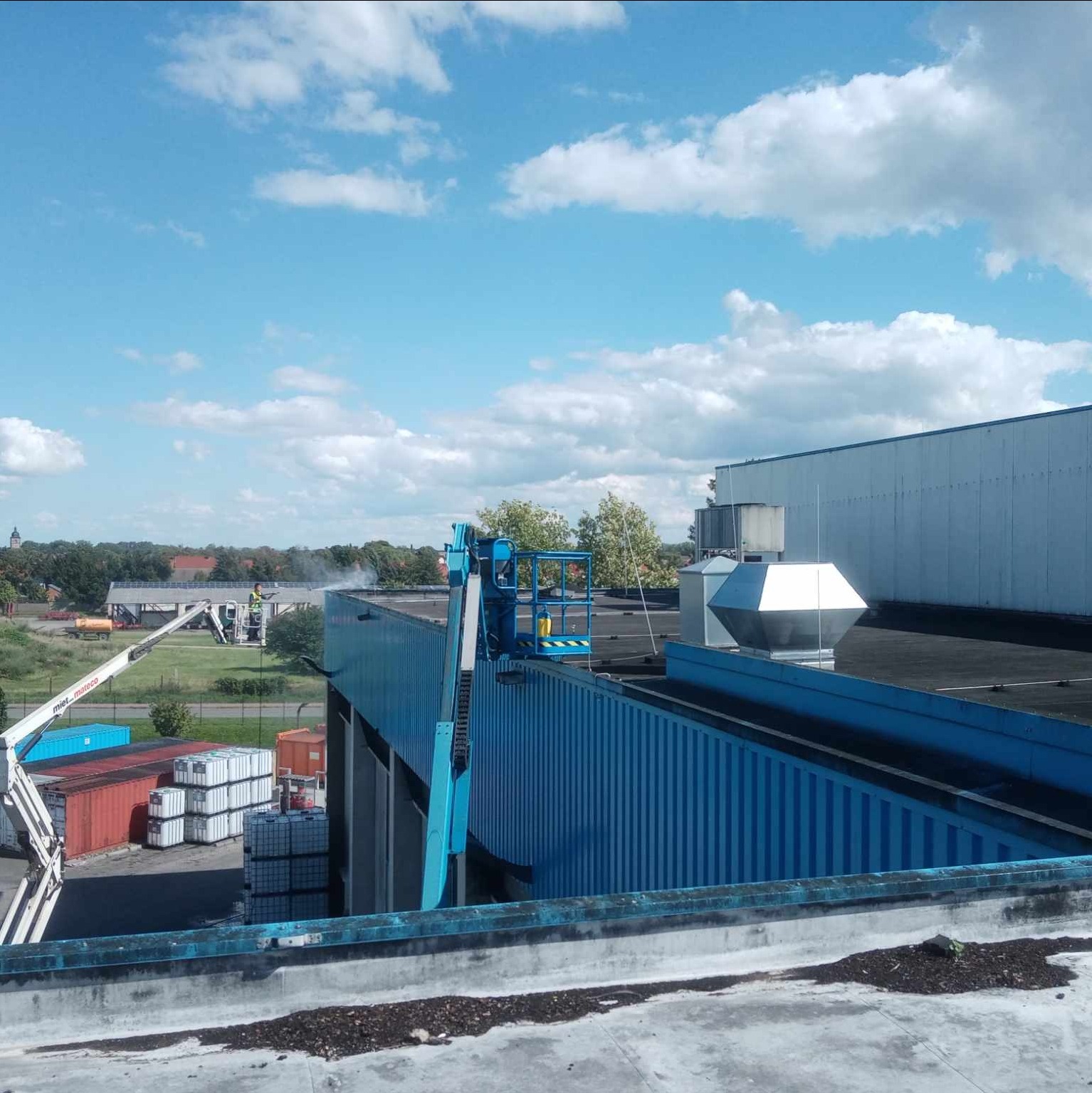 Pracownik w podnośniku koszowym czyści dach budynku przemysłowego za pomocą myjki ciśnieniowej, widoczne kontenery i zabudowania w tle pod błękitnym niebem.