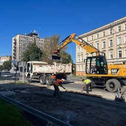 GREGO WŁADYSŁAWA STASZKÓW - Żółta koparka CAT M313D ładuje ziemię do białej ciężarówki na tle miejskiego krajobrazu z budynkami i błękitnym niebem, dwóch robotników wyrównuje teren łopatami