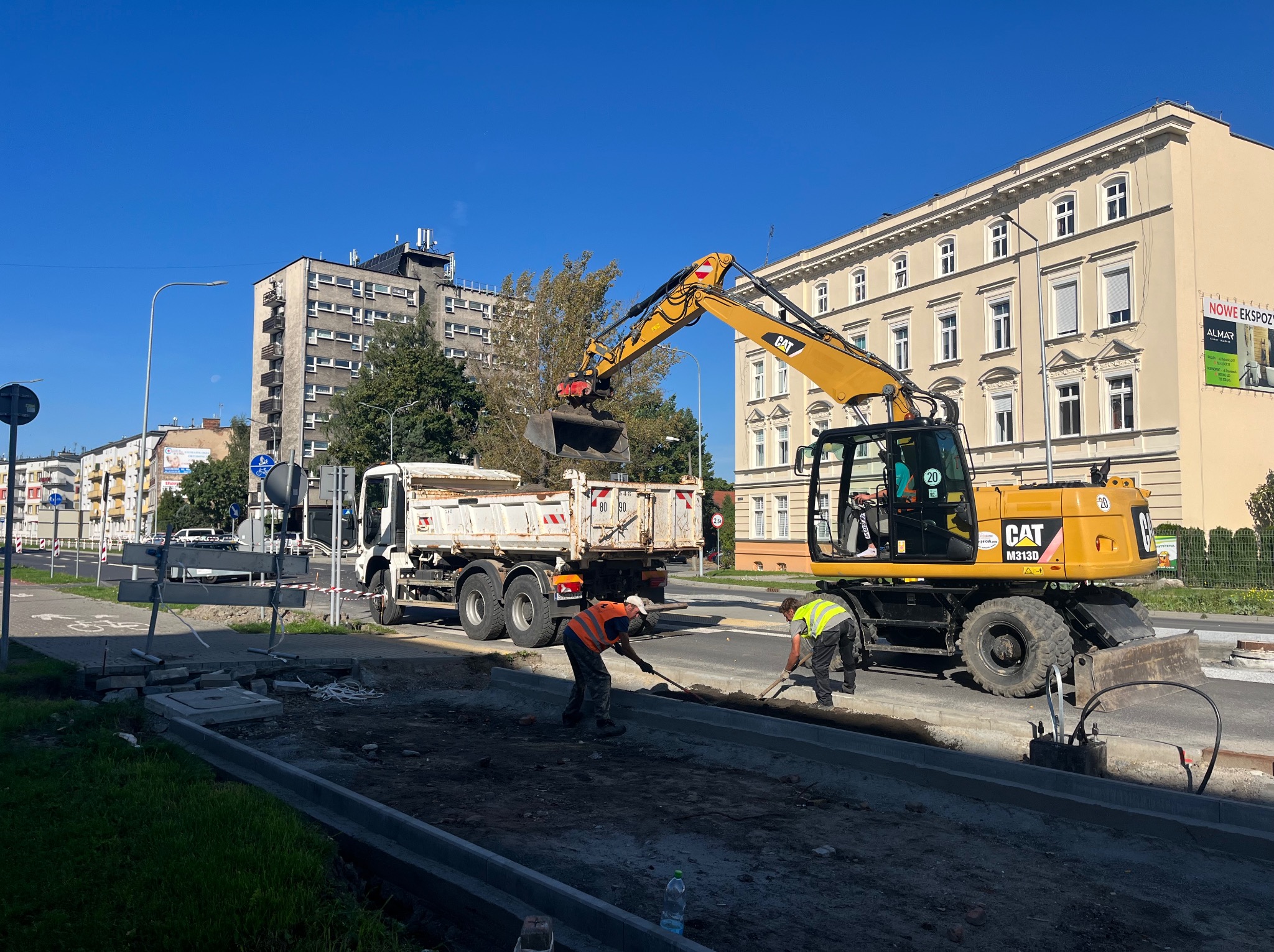 Żółta koparka CAT M313D ładuje ziemię do białej ciężarówki na tle miejskiego krajobrazu z budynkami i błękitnym niebem, dwóch robotników wyrównuje teren łopatami