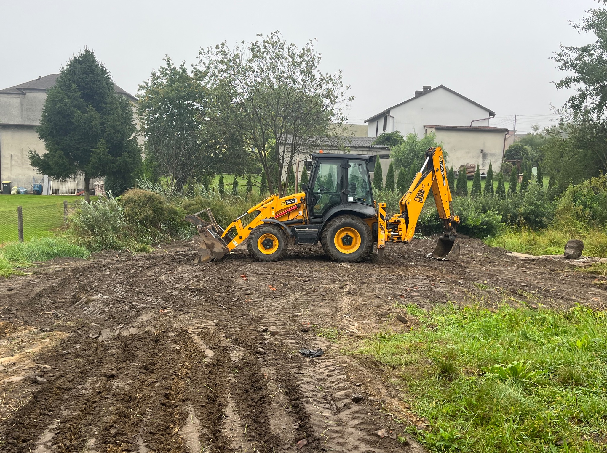 Żółto-czarna koparko-ładowarka JCB na placu budowy z wyraźnymi śladami opon w błotnistej ziemi, w tle zabudowania mieszkalne i zieleń.
