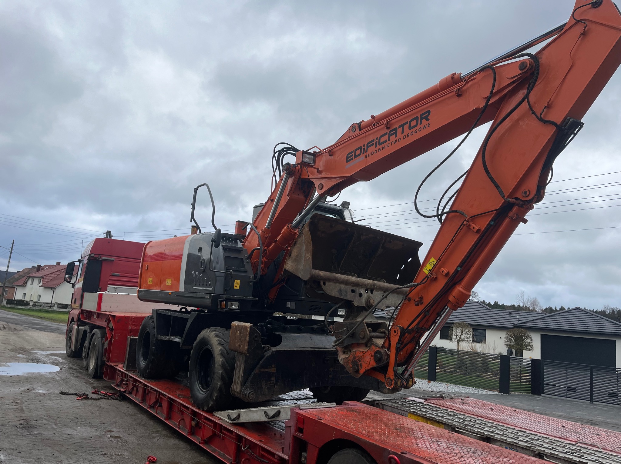 Pomarańczowa koparka kołowa marki Edificator, budownictwo drogowe, transportowana na czerwonej niskopodwoziowej platformie ciężarowej, na tle pochmurnego nieba i zabudowań wiejskich.
