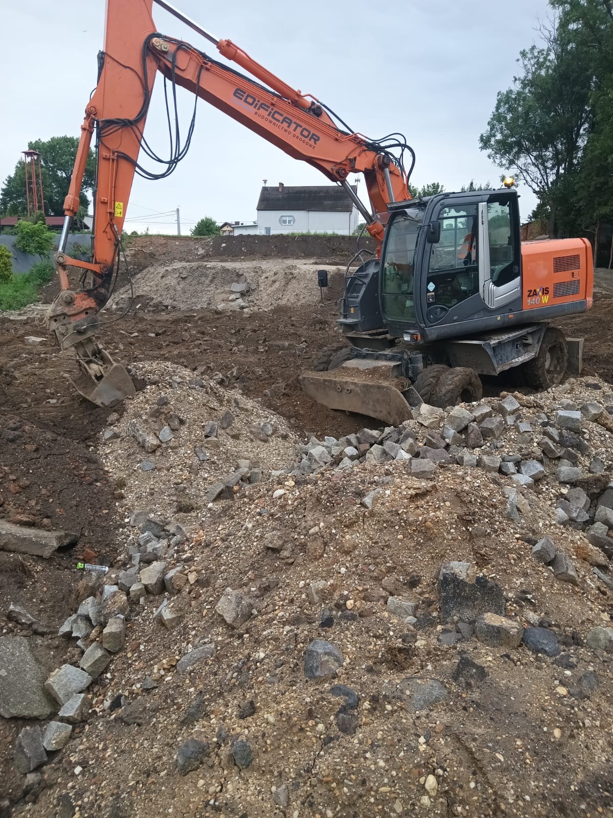 Pomarańczowa koparka firmy Edificator przy wykopie, z hałdą gruzu i ziemi w tle, widoczny budynek mieszkalny i drzewa.