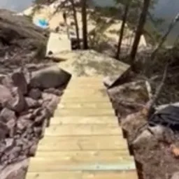 Nowe drewniane schody prowadzące w dół po nierównym, skalistym terenie z drzewami w tle, widok z góry.