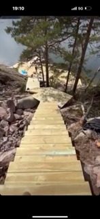 Nowe drewniane schody prowadzące w dół po nierównym, skalistym terenie z drzewami w tle, widok z góry.