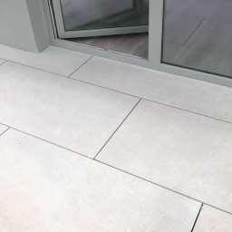 Broker Bud - remonty i wykończenia wnętrz - Jasne, prostokątne płytki podłogowe z ciemnymi fugami, częściowo widoczne szklane drzwi balkonowe z metalową ramą w odcieniach szarości.