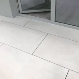 Jasne, prostokątne płytki podłogowe z ciemnymi fugami, częściowo widoczne szklane drzwi balkonowe z metalową ramą w odcieniach szarości.