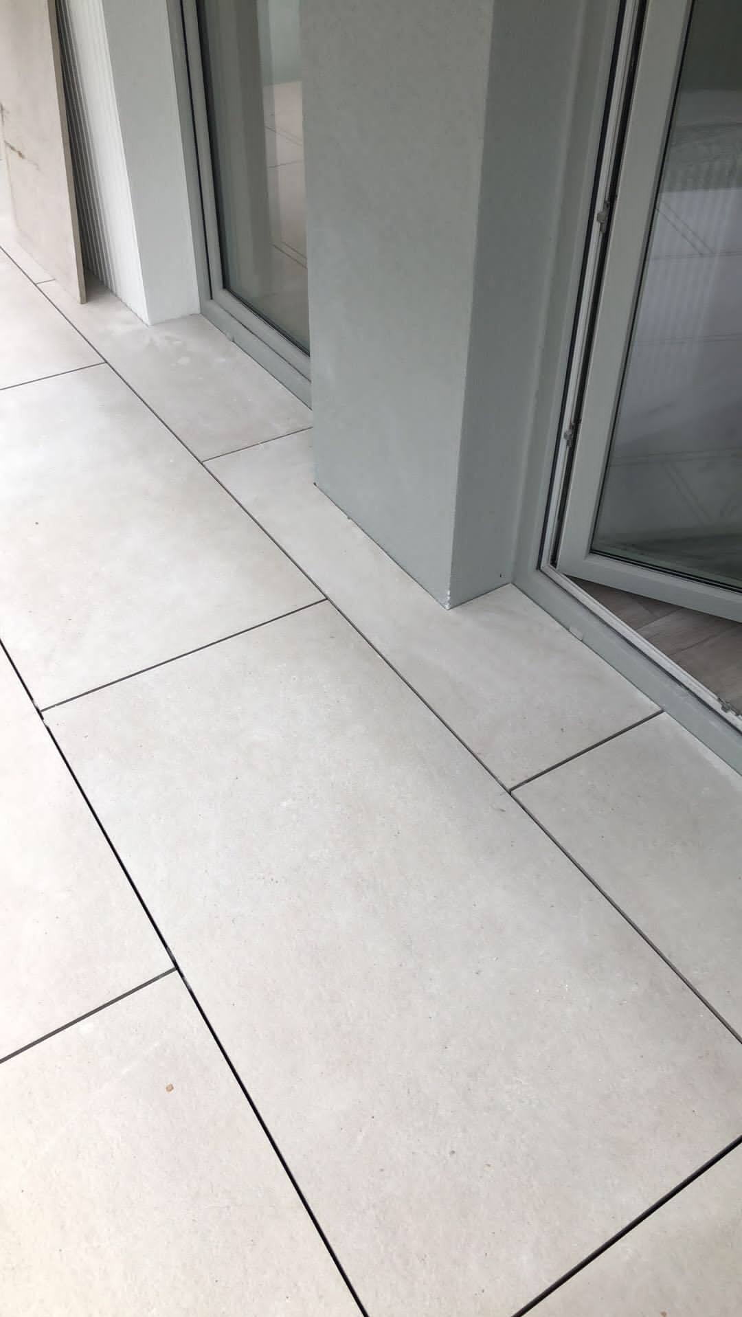 Jasne, matowe płytki podłogowe na balkonie, ułożone w prostokątnym wzorze, przylegające do szarej ramy okiennej. Minimalistyczny design.