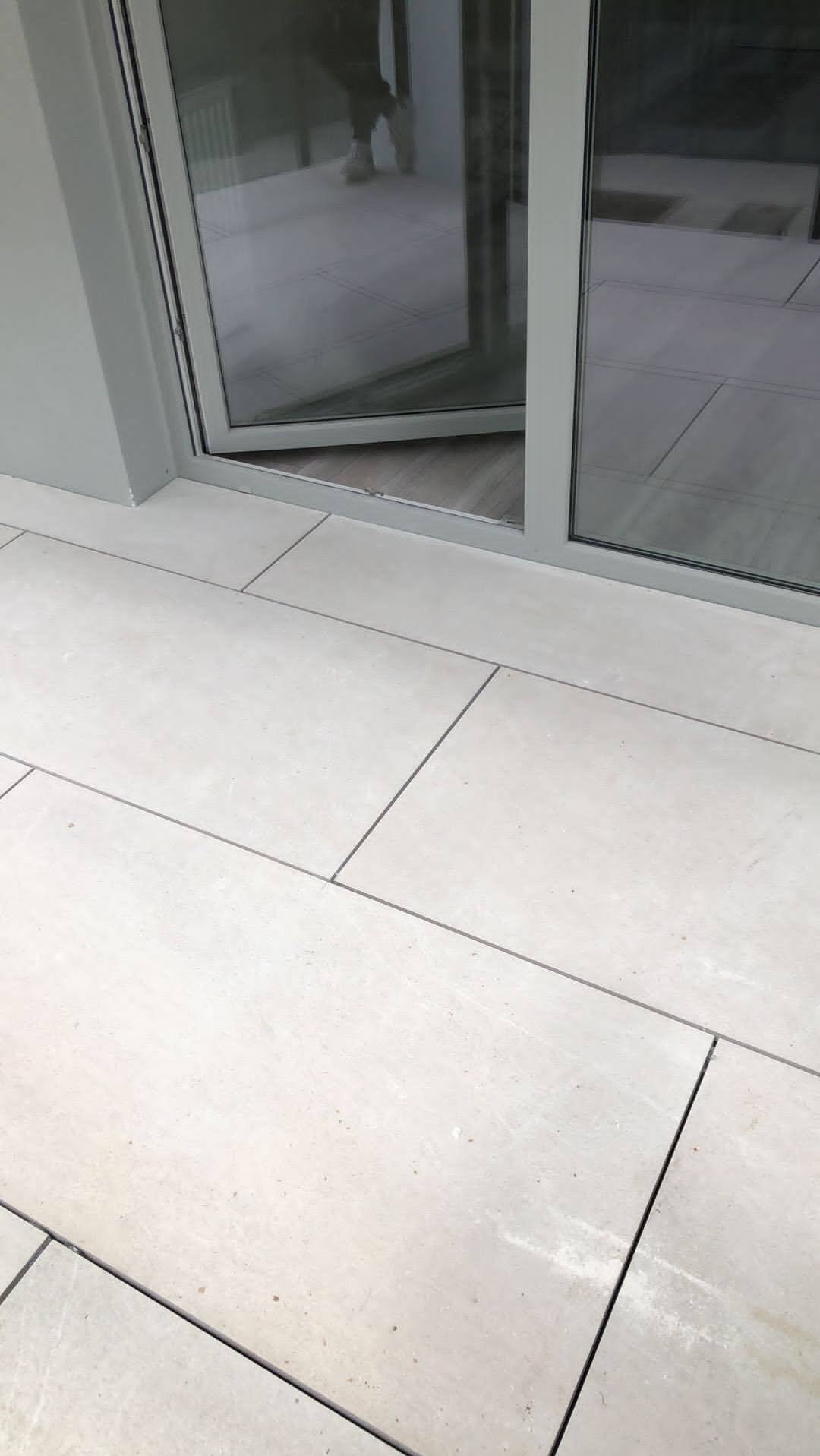 Jasne, prostokątne płytki podłogowe z ciemnymi fugami, częściowo widoczne szklane drzwi balkonowe z metalową ramą w odcieniach szarości.