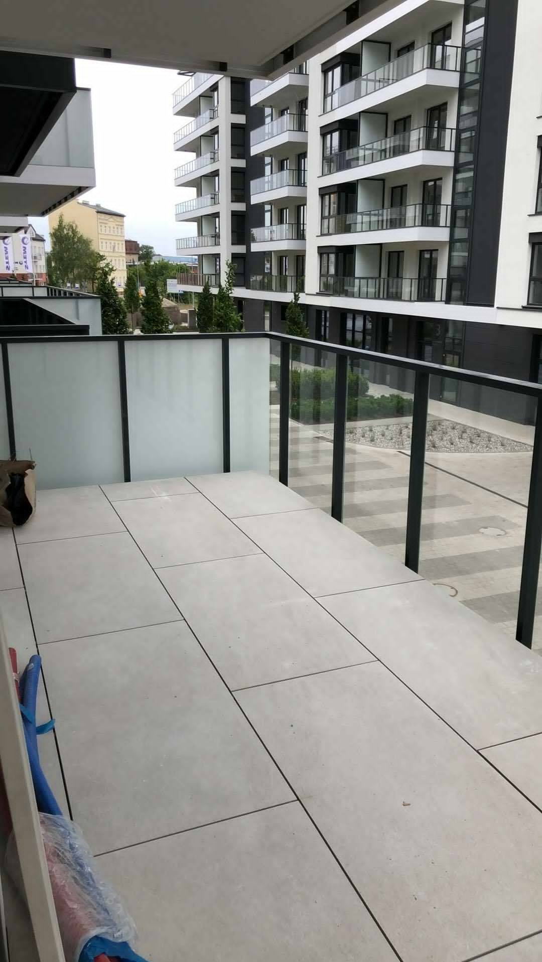 Wykończony balkon w nowoczesnym apartamentowcu. Szare płytki ułożone na podłodze, szklane balustrady. Widok na budynek z wieloma balkonami.