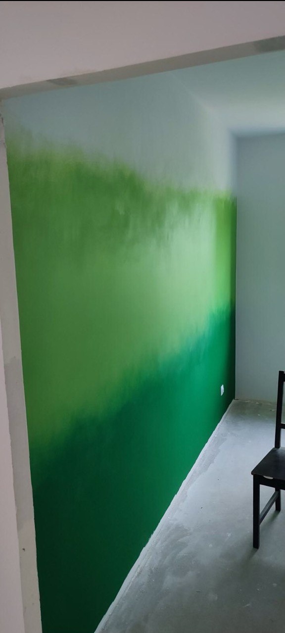 Ściana w pokoju pomalowana farbą w odcieniach zieleni, tworząca efekt ombre. Widoczny fragment podłogi i krzesło. Świeżo pomalowane ściany.