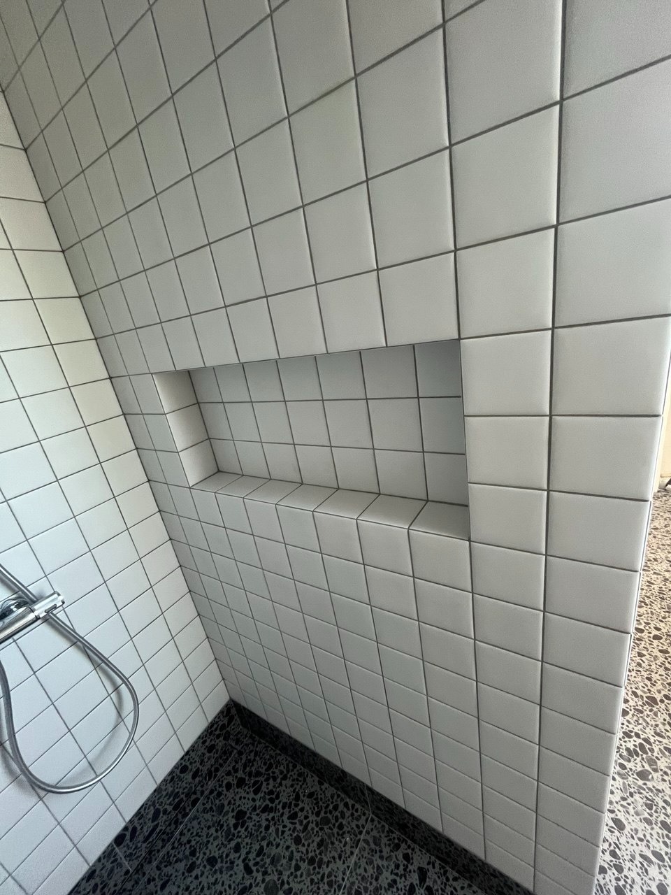 Wykończona kabina prysznicowa z wnęką, wyłożona białymi, kwadratowymi płytkami. Na podłodze czarne lastryko. Widoczny fragment baterii prysznicowej.