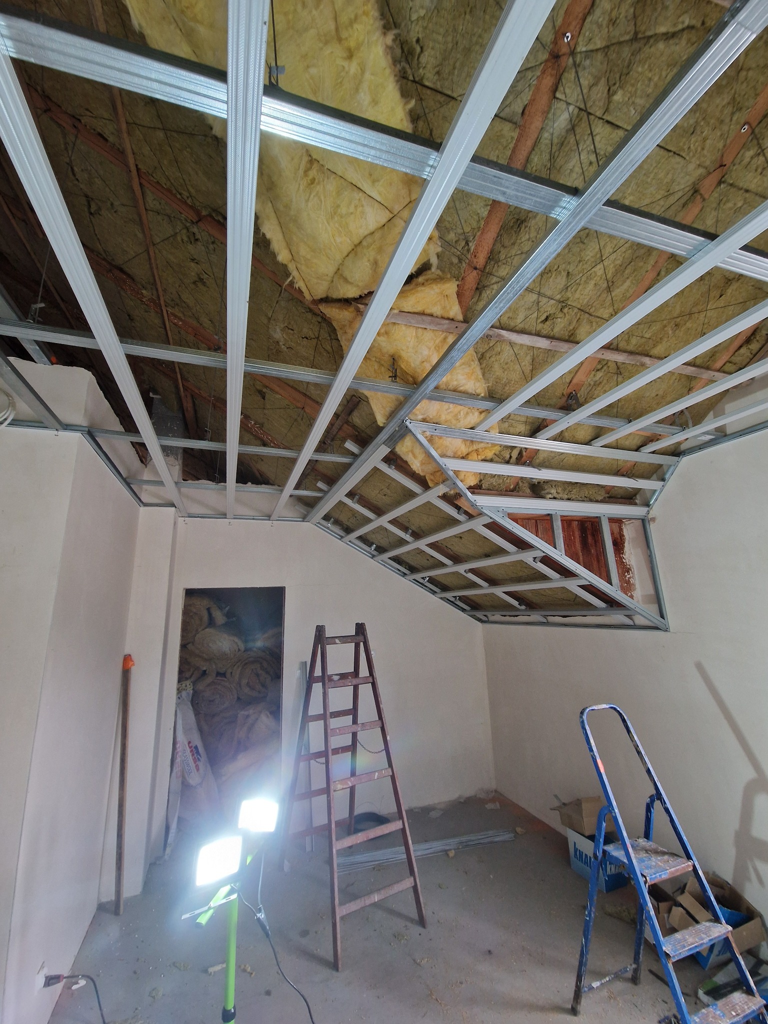 Montaż metalowej konstrukcji sufitu podwieszanego z izolacją z wełny mineralnej, widoczne drabiny i lampa budowlana.