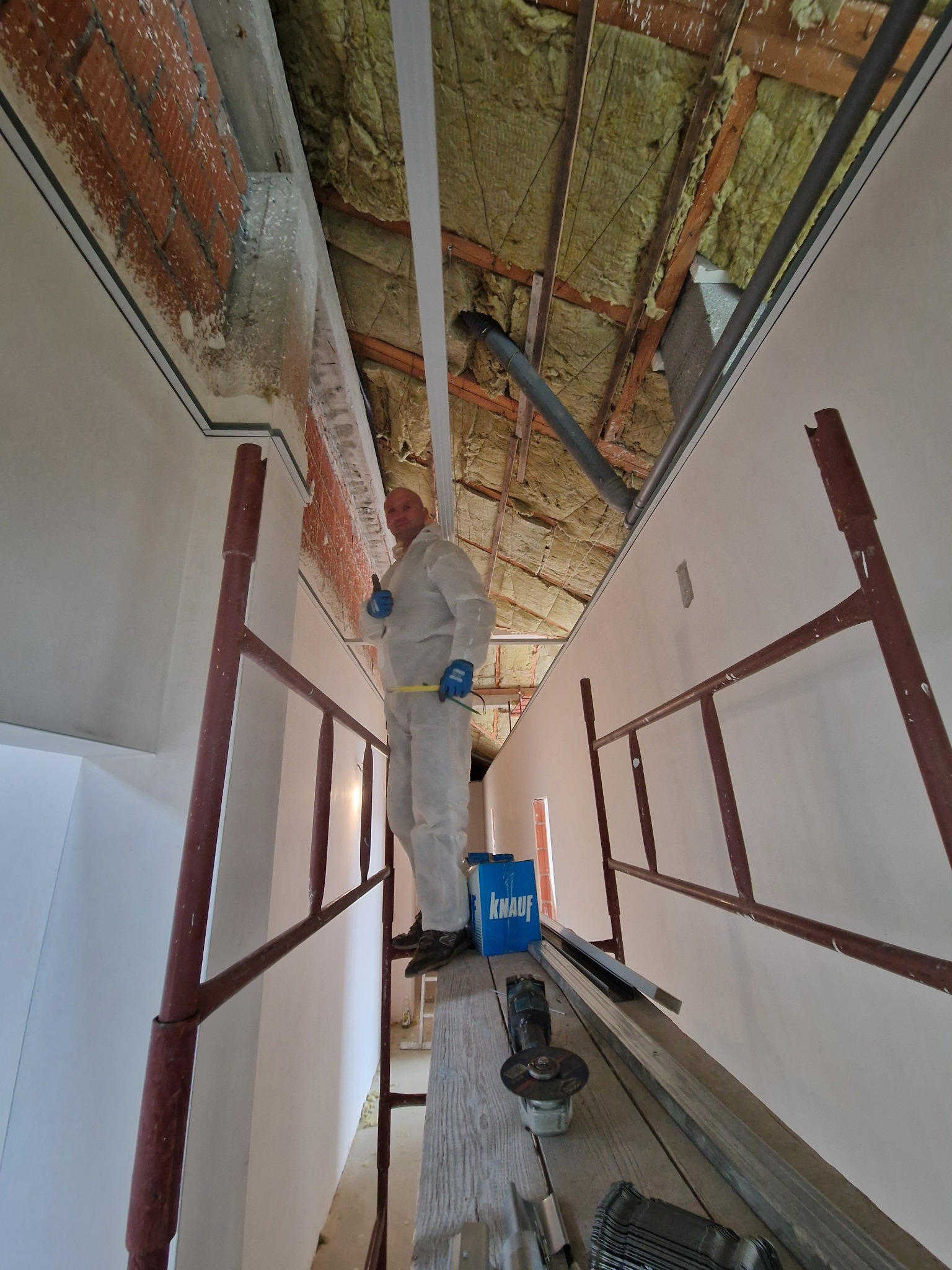 Pracownik w kombinezonie na rusztowaniu, w trakcie montażu profili pod płytę gipsowo-kartonową, widoczne ocieplenie poddasza i narzędzia pracy.