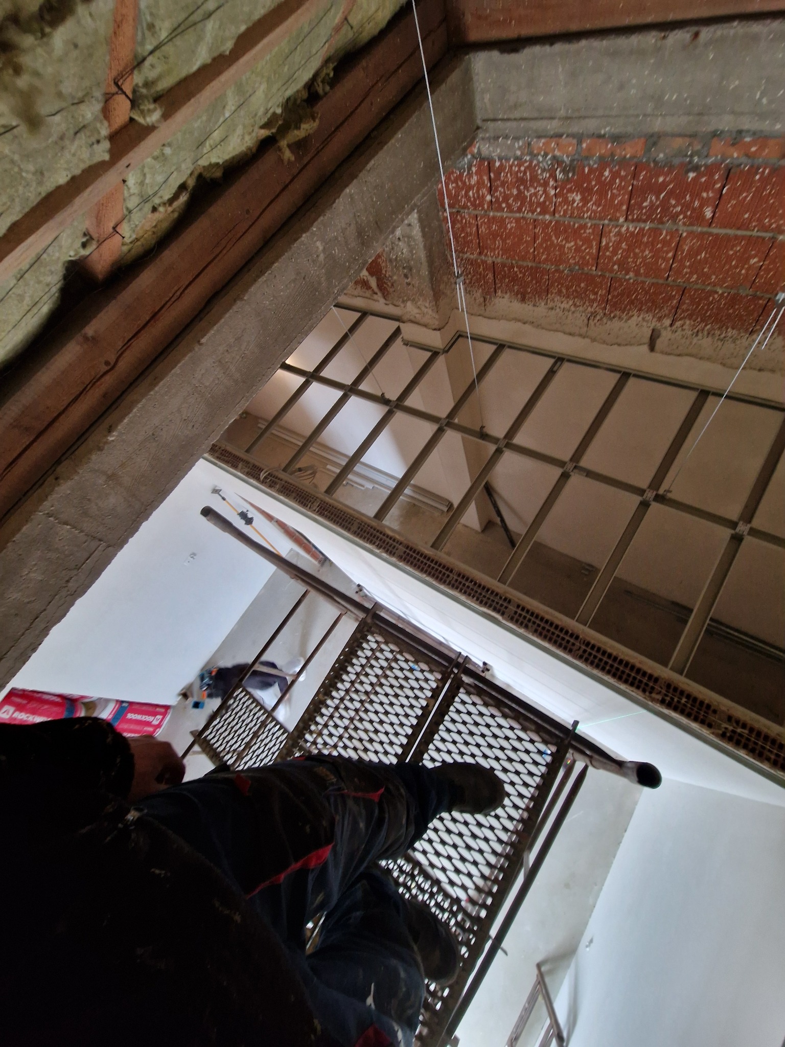 Prace montażowe rusztowania wewnątrz budynku, widoczna izolacja wełną mineralną, surowa cegła i konstrukcja sufitu podwieszanego z płyt karton gips.