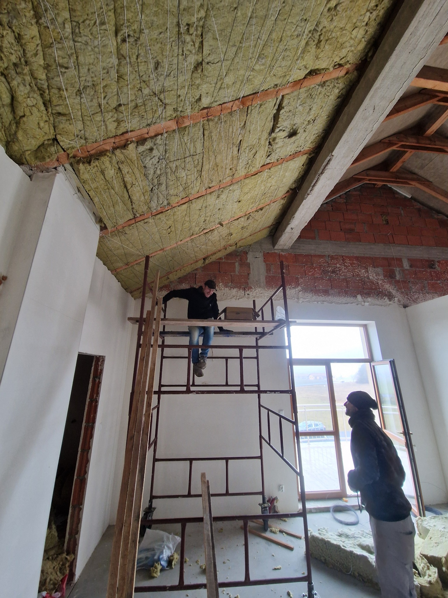 Pracownik na rusztowaniu montuje izolację z wełny mineralnej na poddaszu z widoczną konstrukcją więźby dachowej, drugi pracownik obserwuje postęp prac przy oknie.