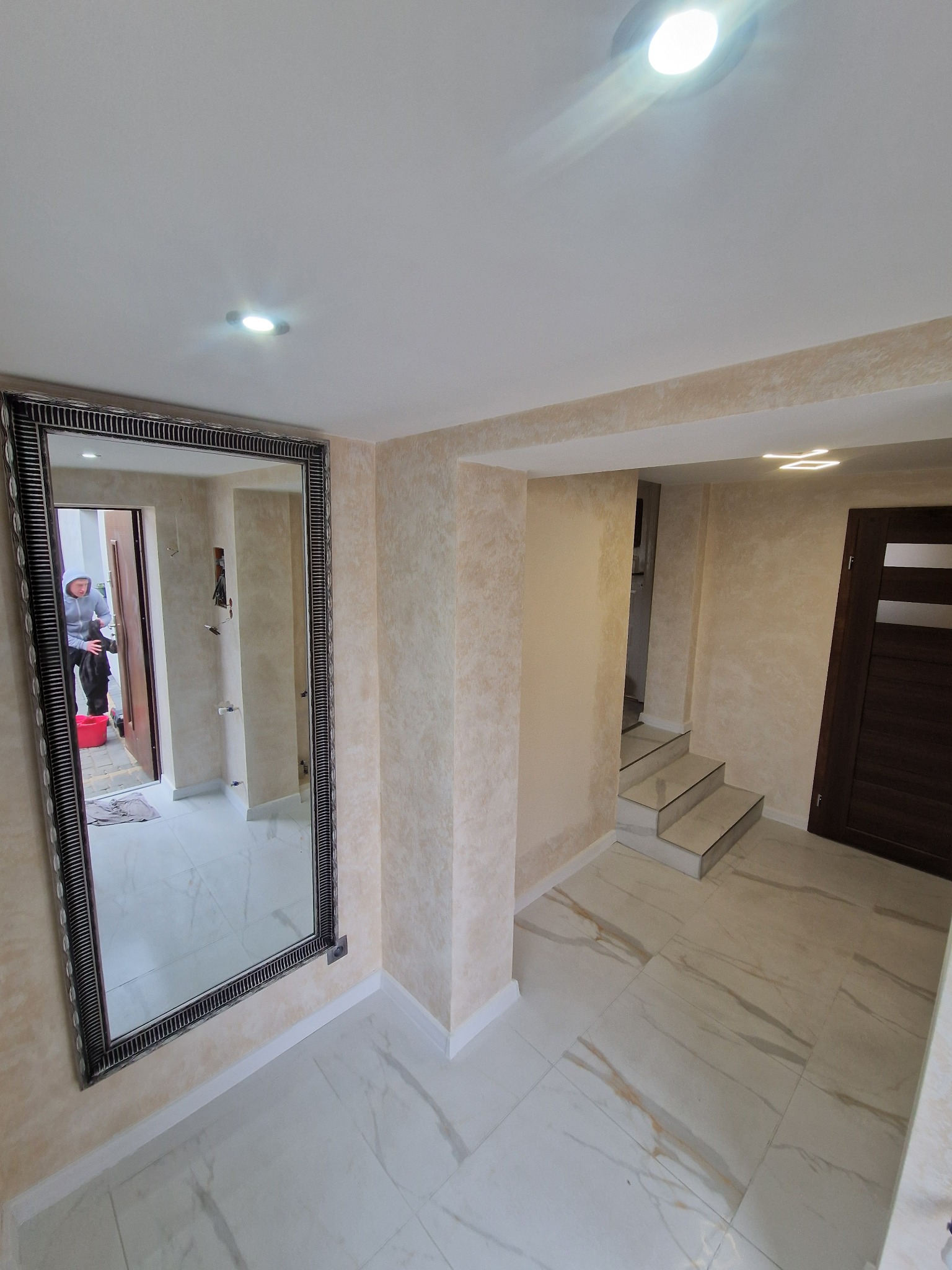 Jasne wnętrze po remoncie: kremowe ściany z dekoracyjną fakturą, biała marmurowa podłoga, duże lustro w ozdobnej ramie odbijające osobę w bluzie, schody z białych płytek prowadzące do kolejnego...