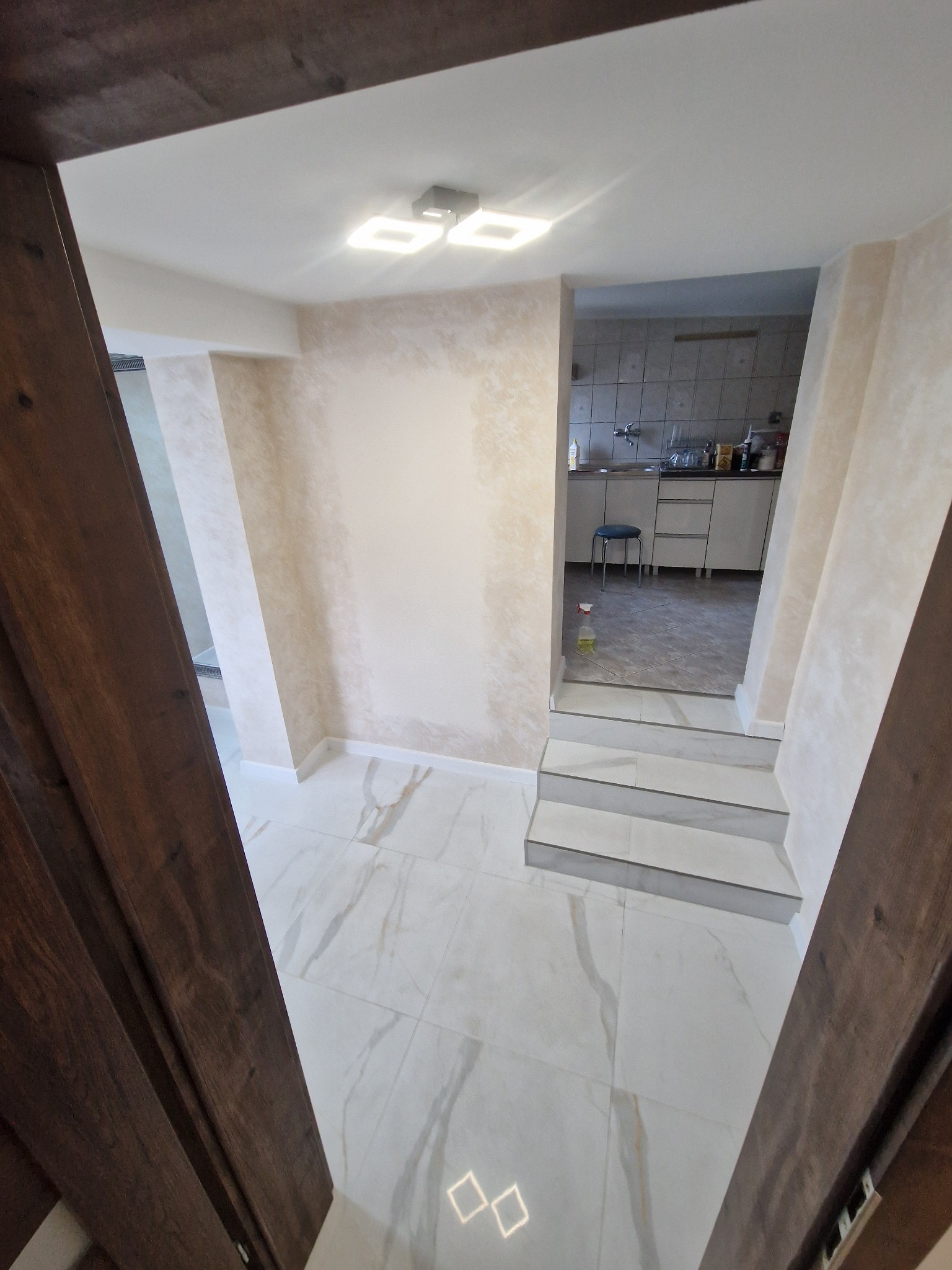 Widok z perspektywy wejścia na fragment wykończonego wnętrza: marmurowe płytki podłogowe, ściany z efektem dekoracyjnym, schody z białymi płytkami prowadzące do kuchni.