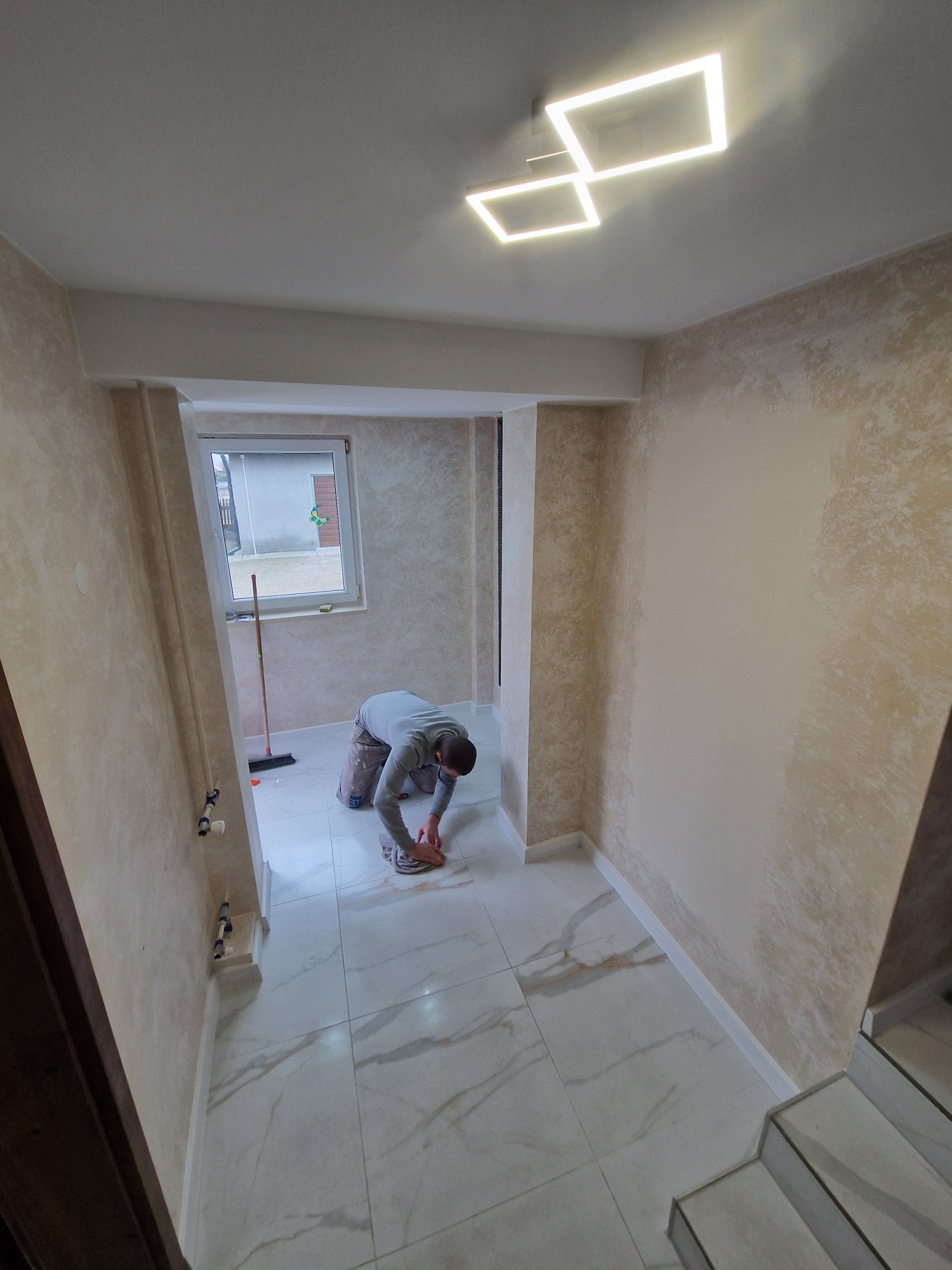 Pracownik na kolanach czyści posadzkę z płytek marmuropodobnych w korytarzu po zakończonych pracach wykończeniowych, widoczne świeżo pomalowane ściany z efektem dekoracyjnym, nowoczesna lampa...